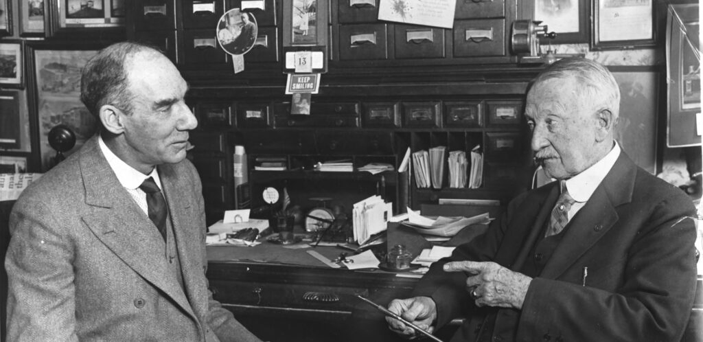 Black and white photograph of two men talking in front of a collection of objects.