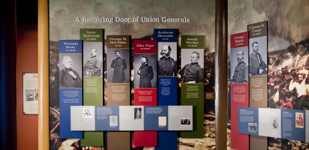 A display with eight blue, green, red, and brown panels, each bearing a black and white photograph of a Union general. Behind the panels is a painting of soldiers engaged in combat.