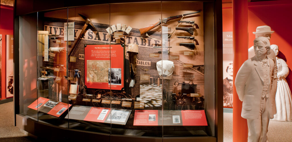 A museum display featuring artifacts including five guns, a white hood, a pick ax and five small portraits. To the right of the display are three white life-sized statues representing some of the conspirators in the Lincoln assassination.