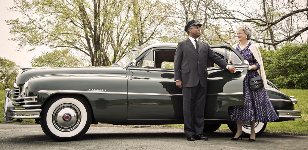 Actors portraying Miss Daisy Werthan and her chauffeur Hoke Colburn. Hoke is opening the backdoor of a 1948 Packard for Daisy. They are smiling at each other.