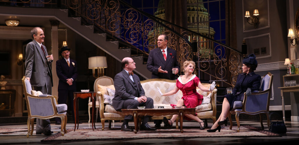 On a set designed to look like a fancy hotel living room in the 1950s, a woman in a red dress and a man in a three-piece suit sit on a sofa. Four others stand around them.