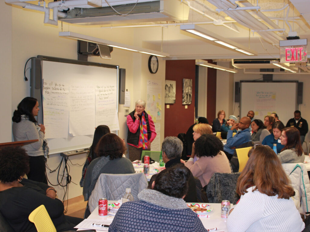 20 teachers are seated at tables of four. They listen to a speaker who is standing near a whiteboard. On the white board are several big sheets of paper, each with a different discussion prompt (illegible from the camera’s vantage at the back of the room.