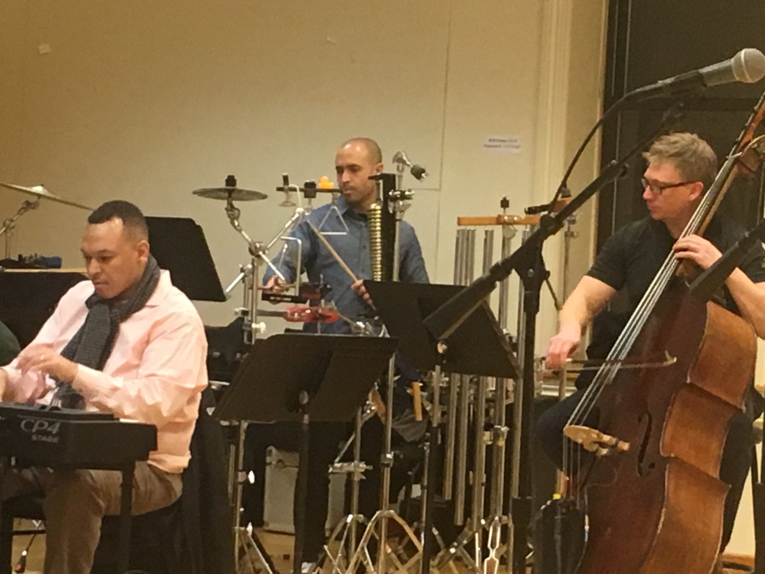 Three musicians sit behind drums and keyboards in a theatre rehearsal studio.