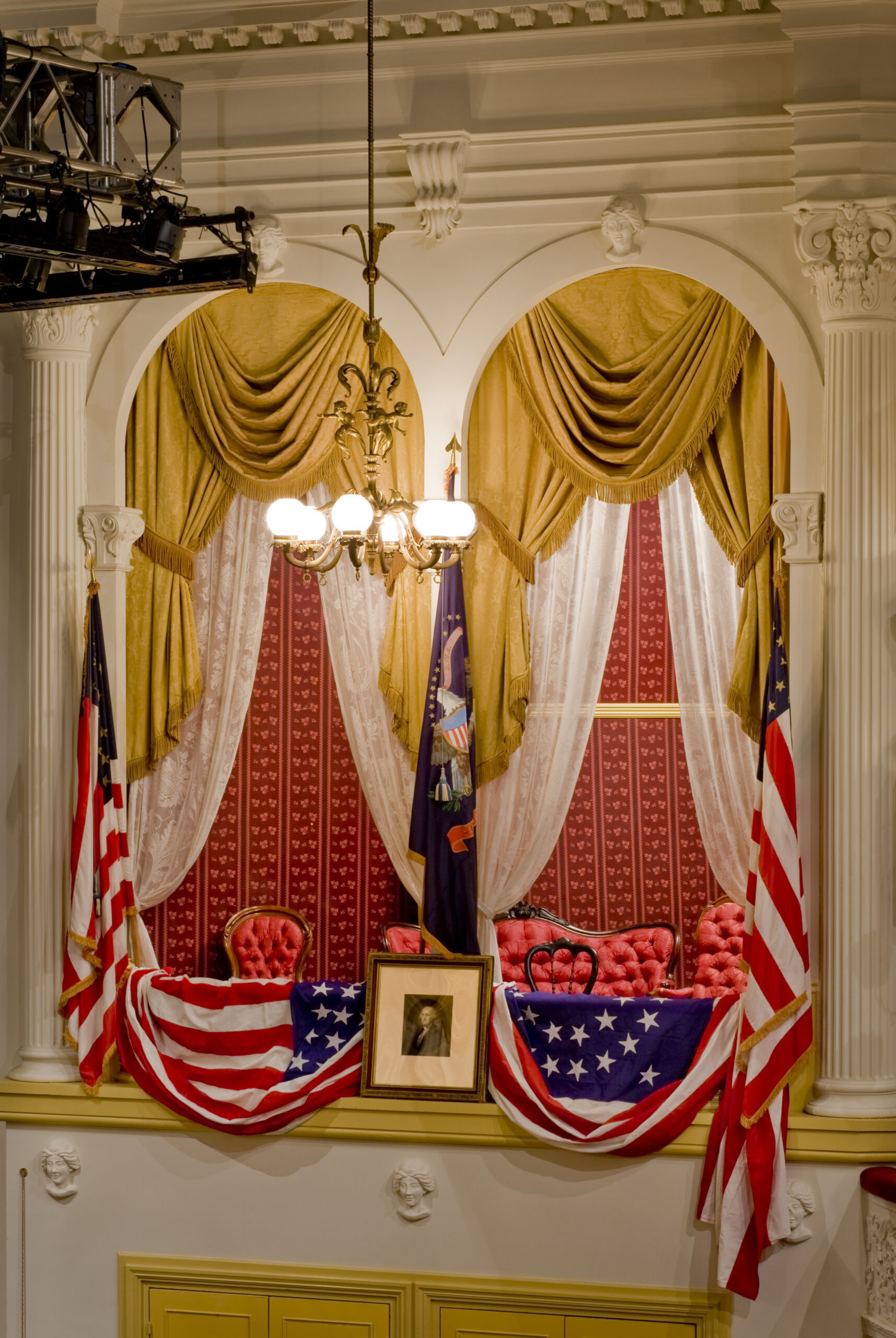 The President’s Box at Ford’s Theatre with American flags on either side, a picture of George Washington in the center and American flag bunting draped over the box. The interior of the box has red patterned wallpaper and red upholstered chairs and a couch. The box is framed by gold curtains with white lace curtains underneath them.