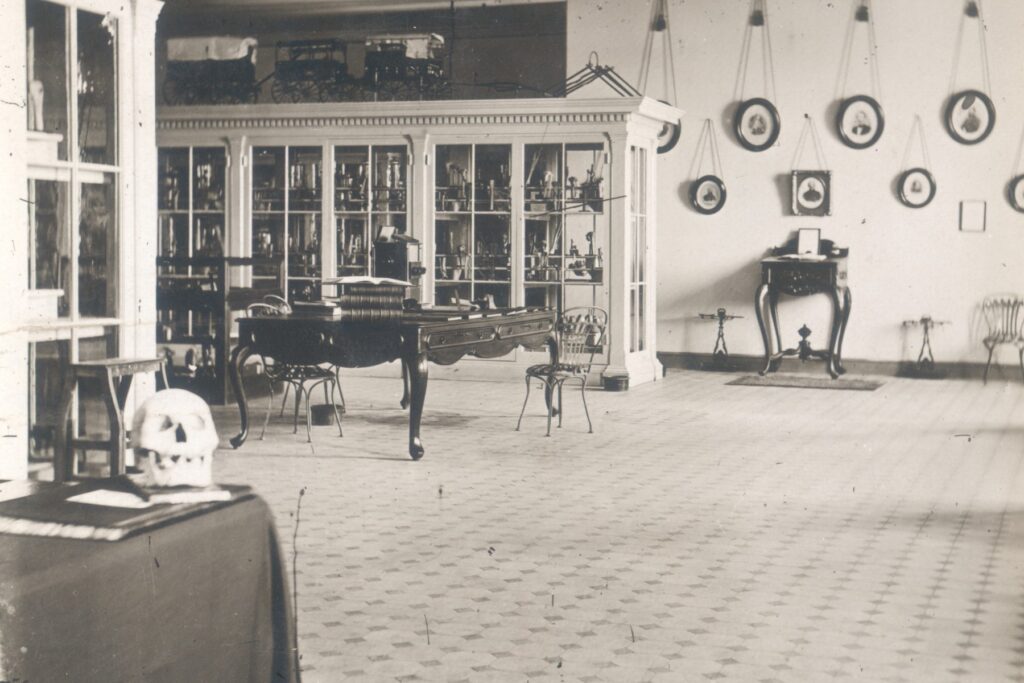 In 1867 the Army Medical Museum opened at Ford's Theatre. This black and white image shows a museum room with portraits hanging on a far wall and several central glass-windowed displace cases. The foreground of the photo features a table with a human skull on it.