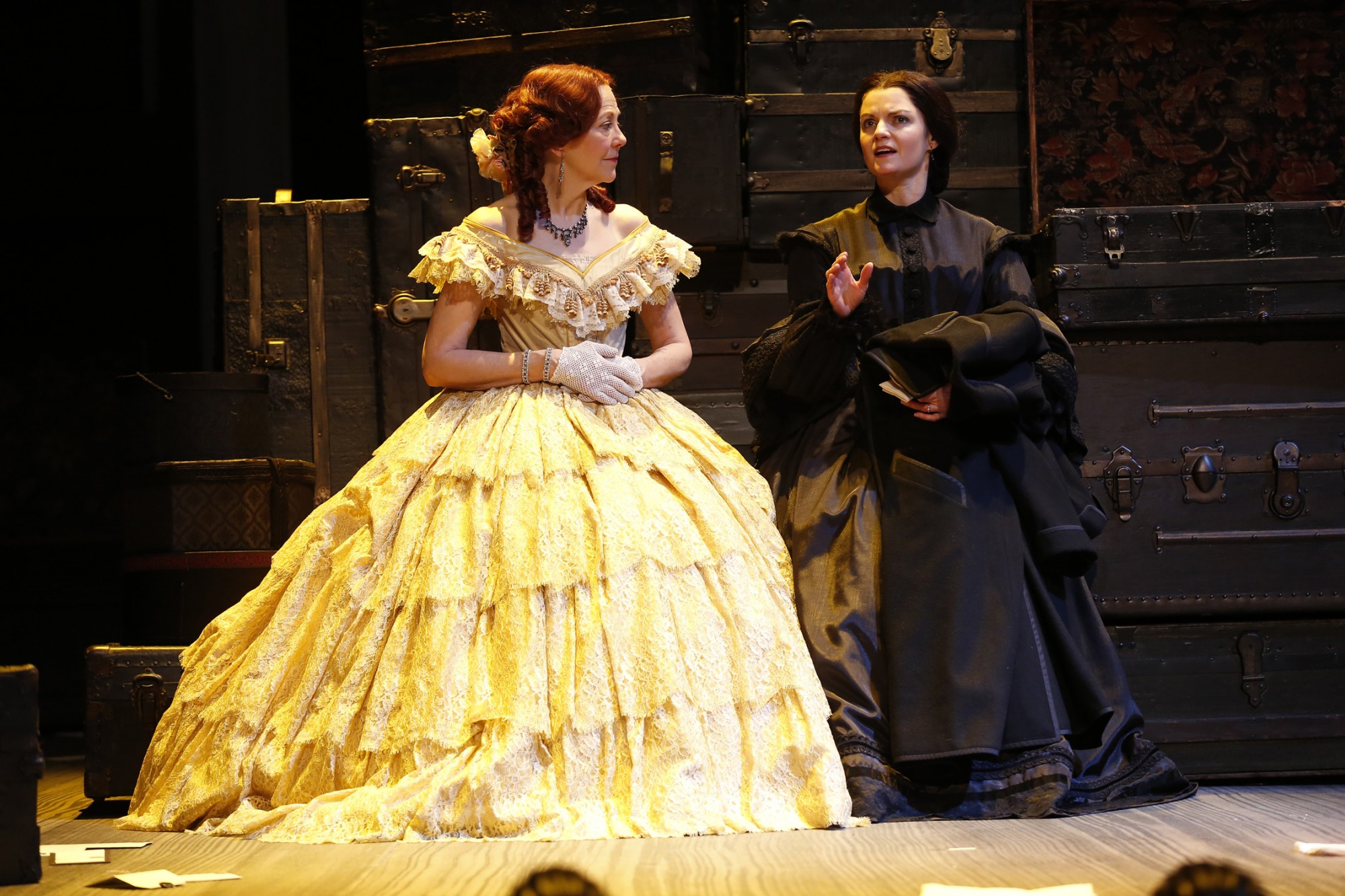 An actress wearing a yellow Victorian gown and white gloves sits beside an actress wearing a black Victorian mourning gown.