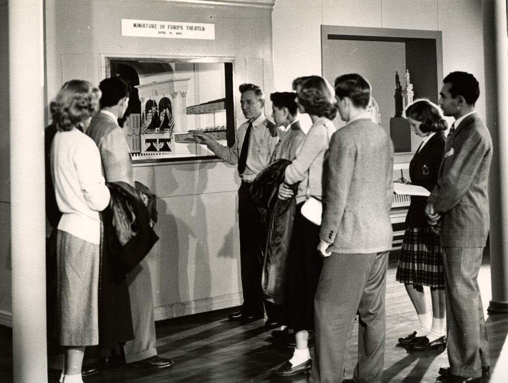 Black and white photo of a group of people looking at a diorama of Ford's Theatre.