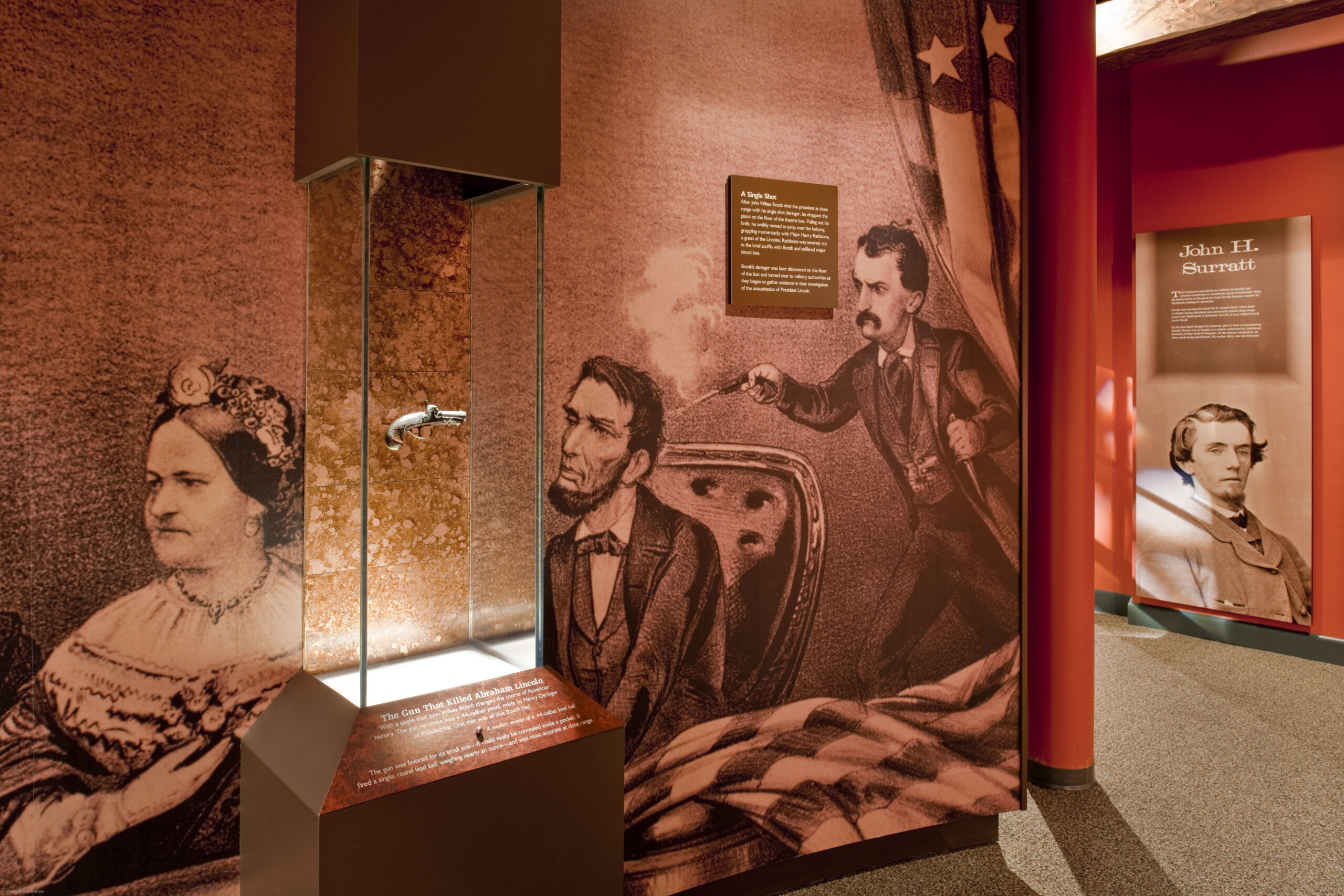 In the middle of a tall glass display case, a small silver gun appears to float in mid-air. Behind the case is a wall-sized drawing of John Wilkes Booth shooting Abraham Lincoln.