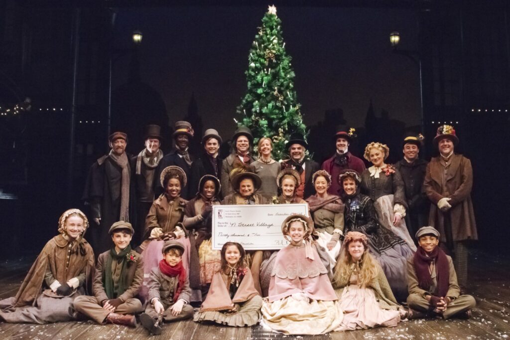 A group of actors dressed in Victorian outfits pose in front of a christmas tree in three rows. The second row holds a large check.