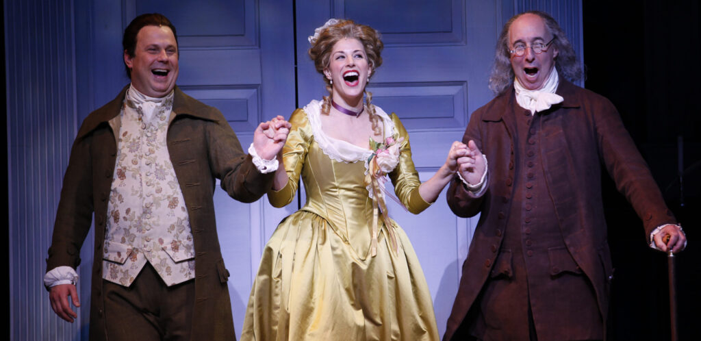 Actors portraying John Adams and Benjamin Franklin each hold one hand of a woman in a yellow dress and sing.