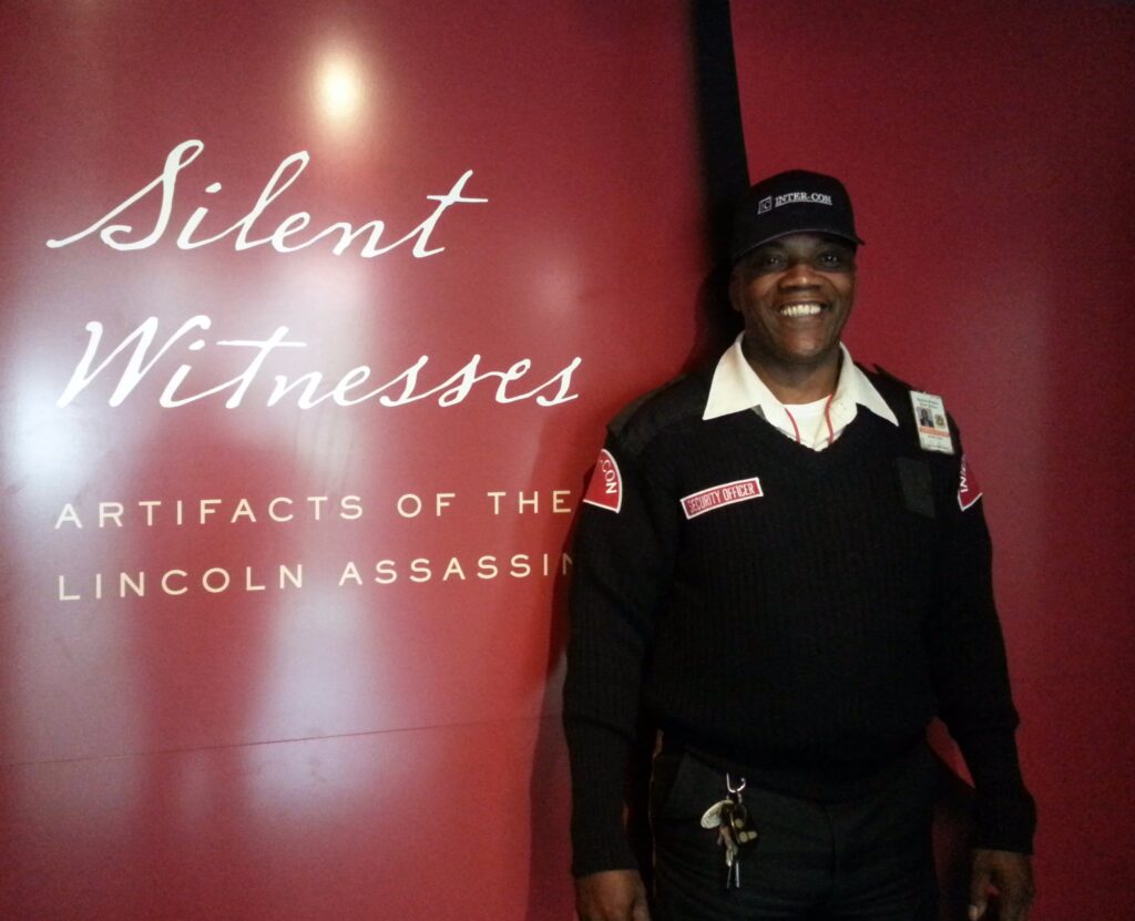 Photograph of a man in a guard uniform standing in front of a wall panel that reads "Silent Witnesses: Artifacts of the Lincoln Assassination."