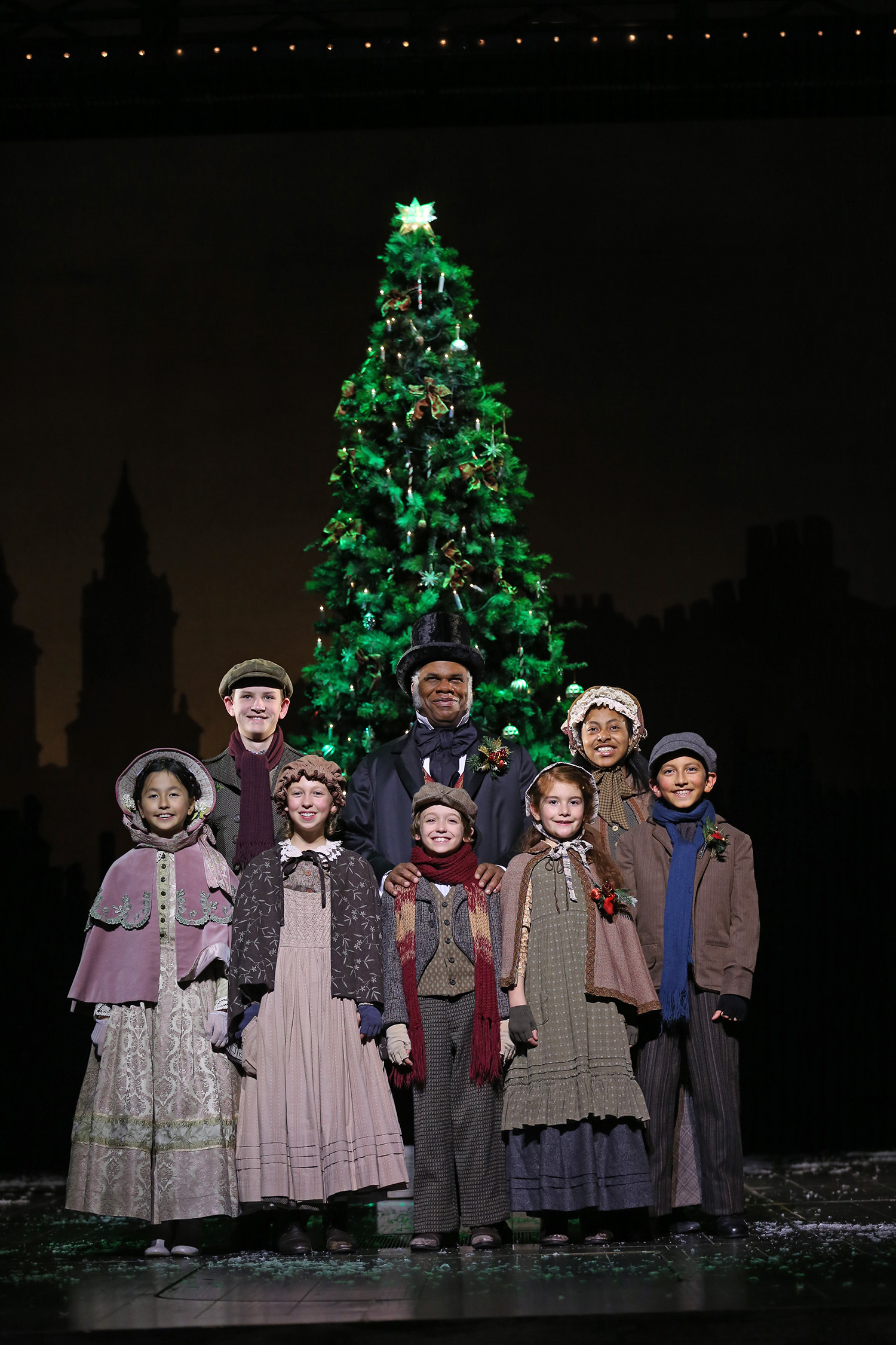 A smiling man in Victorian-style clothes and a top hat stands with a group of children, also in Victorian-style clothing, in front a large Christmas tree.