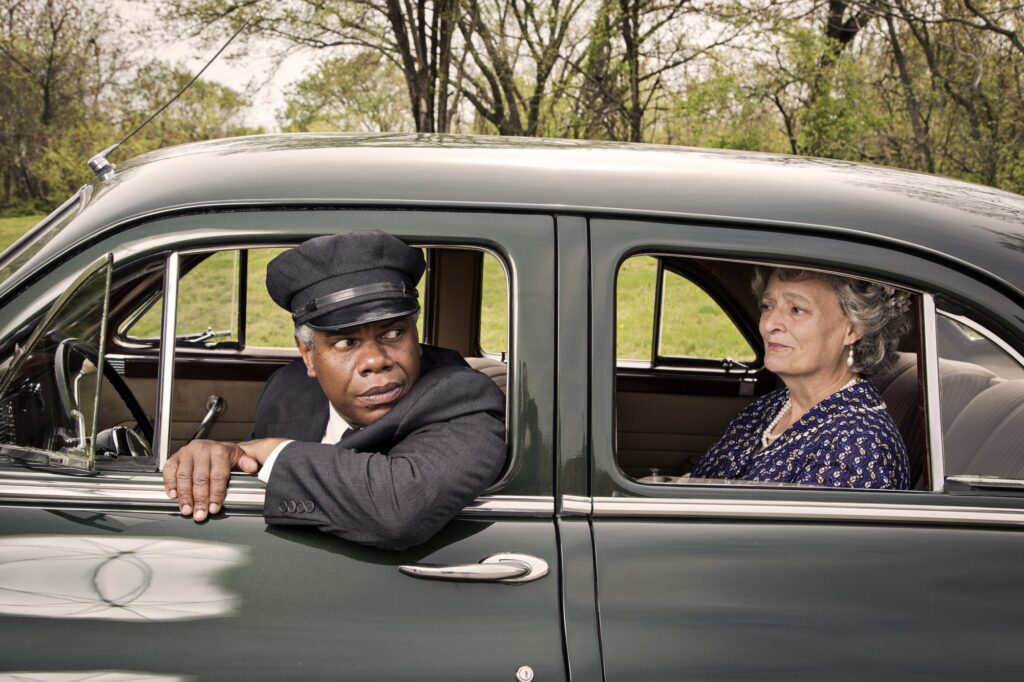 Actors portraying Miss Daisy Werthan and her chauffeur Hoke Colburn. They are seated in a 1948 Packard. Hoke leans out the window of the driver’s seat and wears a concerned expression. Daisy sits in the pack and surveys Hoke.