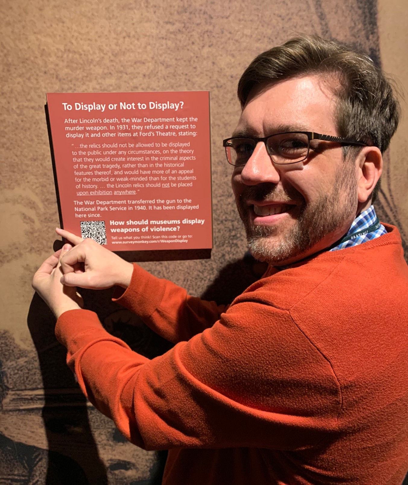 Man smiles while hanging a square orange sign on the Ford’s Theatre Museum wall. The sign reads “To display or not to display?” with information about the National Park Service’s acquisition of John Wilkes Booth’s pistol.