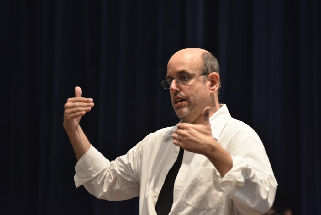 A man in a white shirt and tie gestures and speaks in front of a black background.