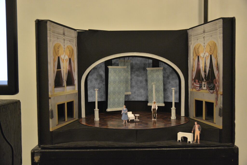 A model of the set for the Ford’s Theatre production of “Driving Miss Daisy.” A series of blue flats hang in the background. There are three white columns and a white chair on stage. Cutout figures representing the characters of Hoke, Daisy and Boolie are also on stage.