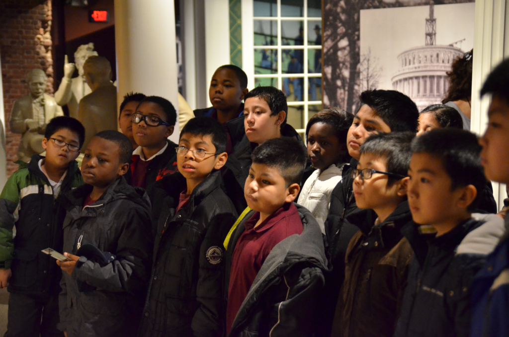 Young children stand together and look at a museum exhibit.