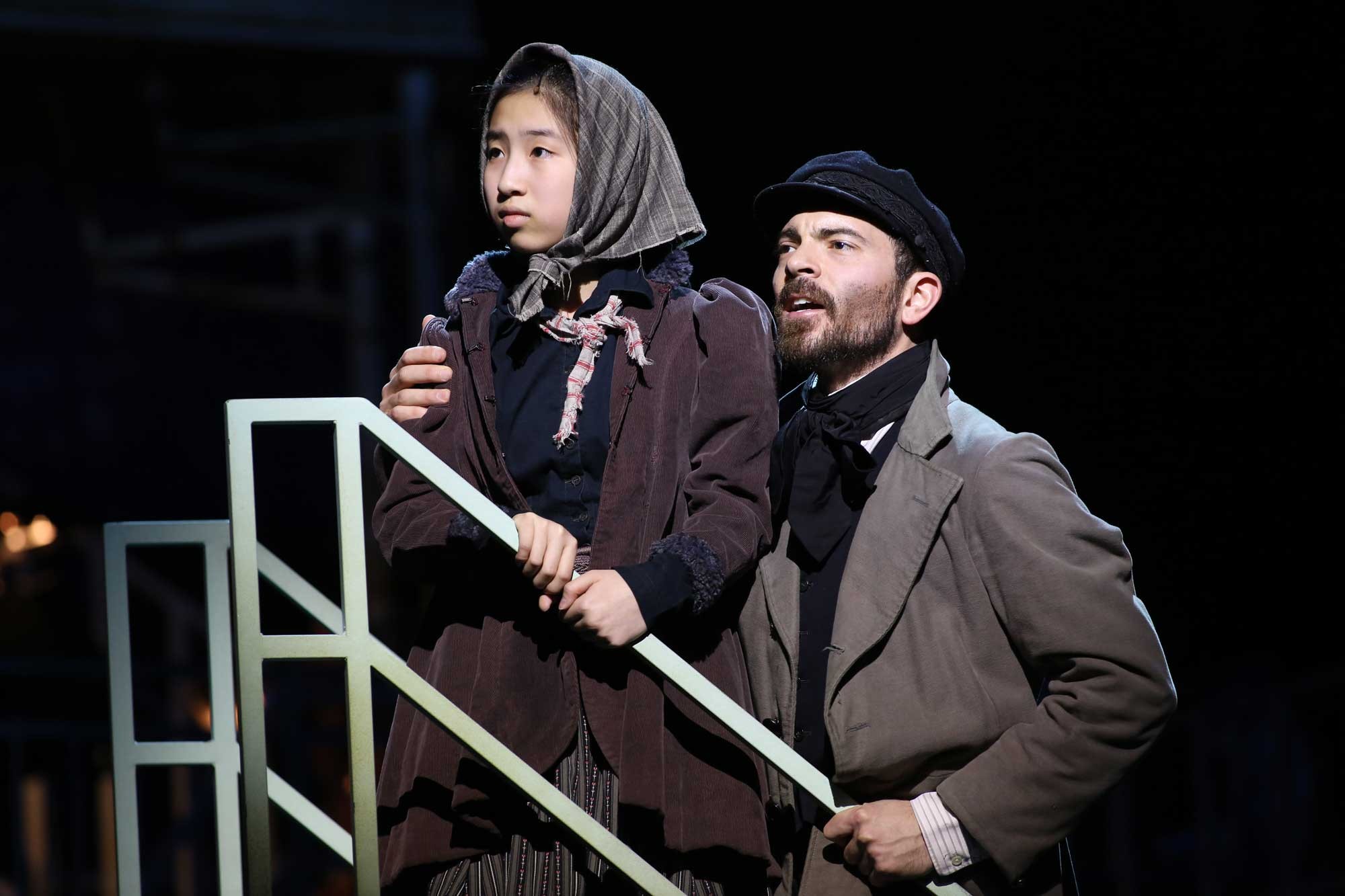 Photo of two actors: one man (Jonathan Atkinson) and one young girl (Dulcey Pham). They are dressed as Jewish immigrants in modest clothing. The young girl wears a headscarf. They are standing near a retail kiosk where the man sells silhouette portraits. Image is from the Ford's production of "Ragtime."  