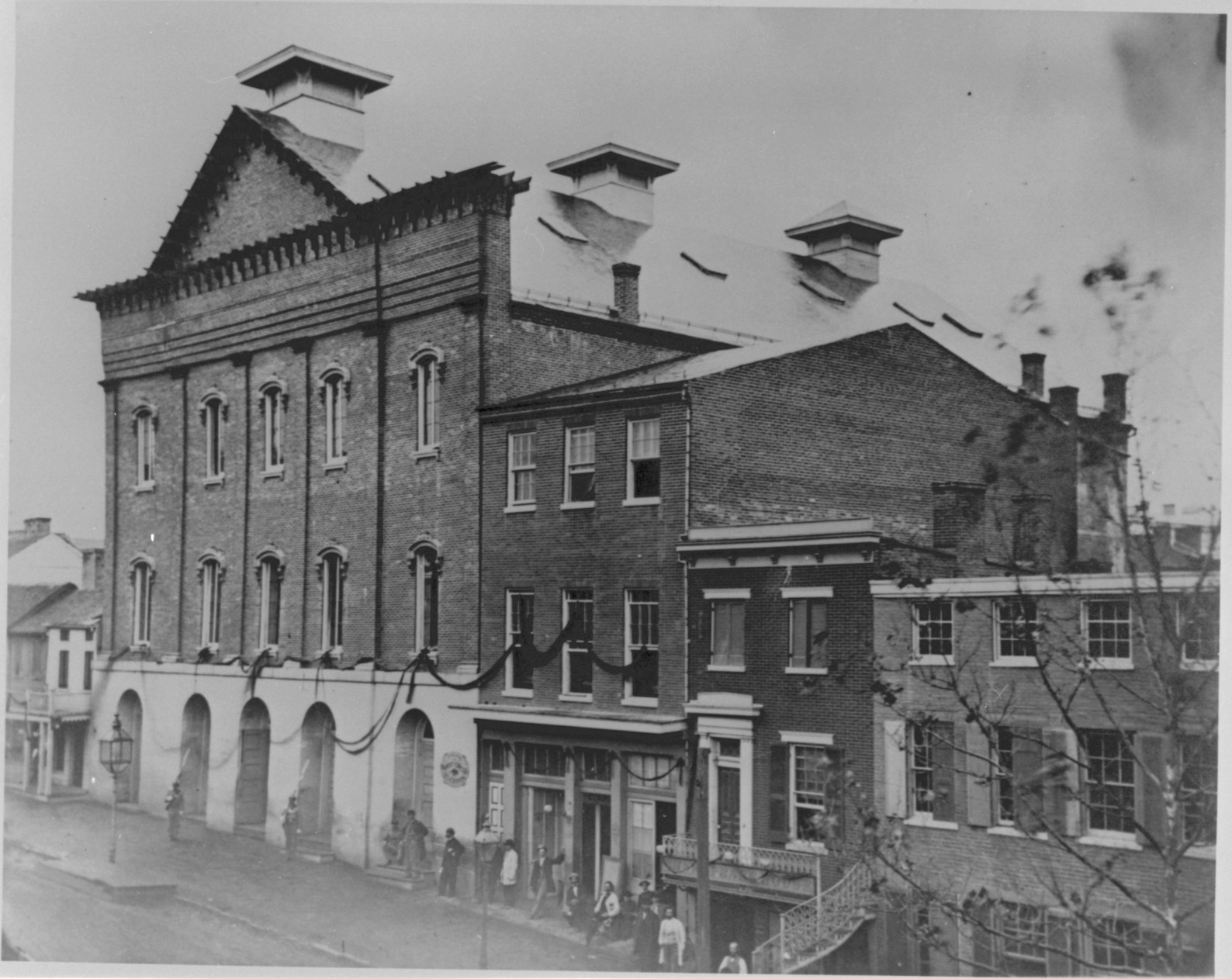 1865 black and white photograph of the exterior of Ford's Theatre in D.C. The image shows drapes of black fabric hanging horizontally between each window in mourning for the death of Abraham Lincoln.