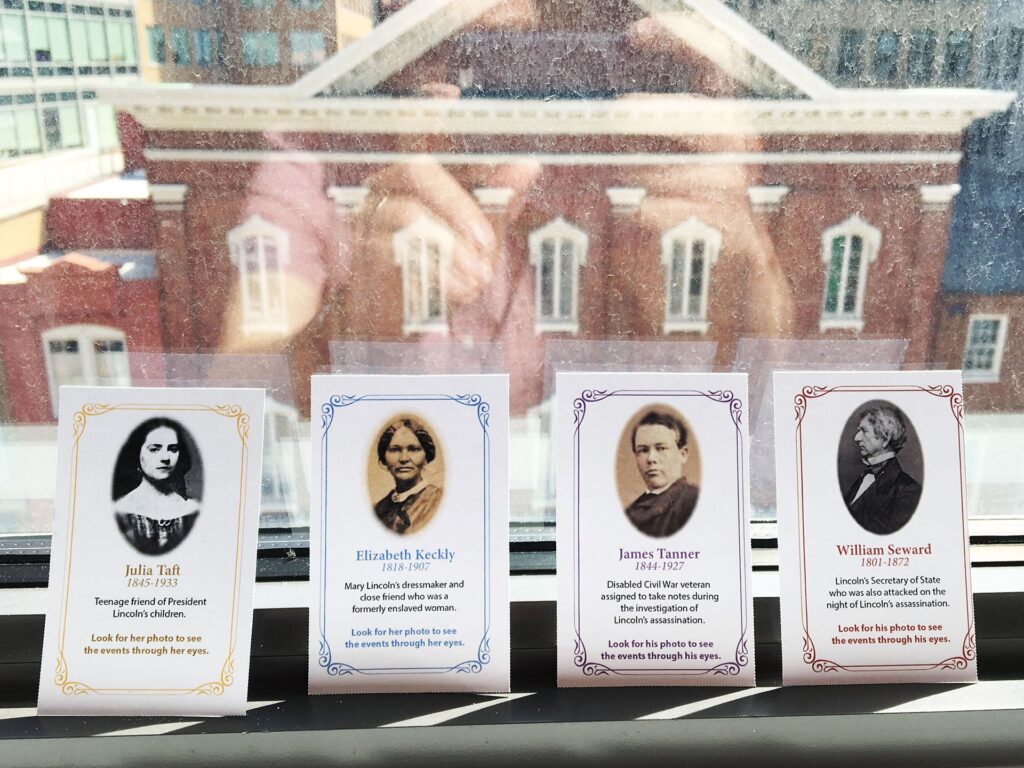 Four cards with pictures of 19th century figures lean against a window. Through the window Ford's Theatre is visible.