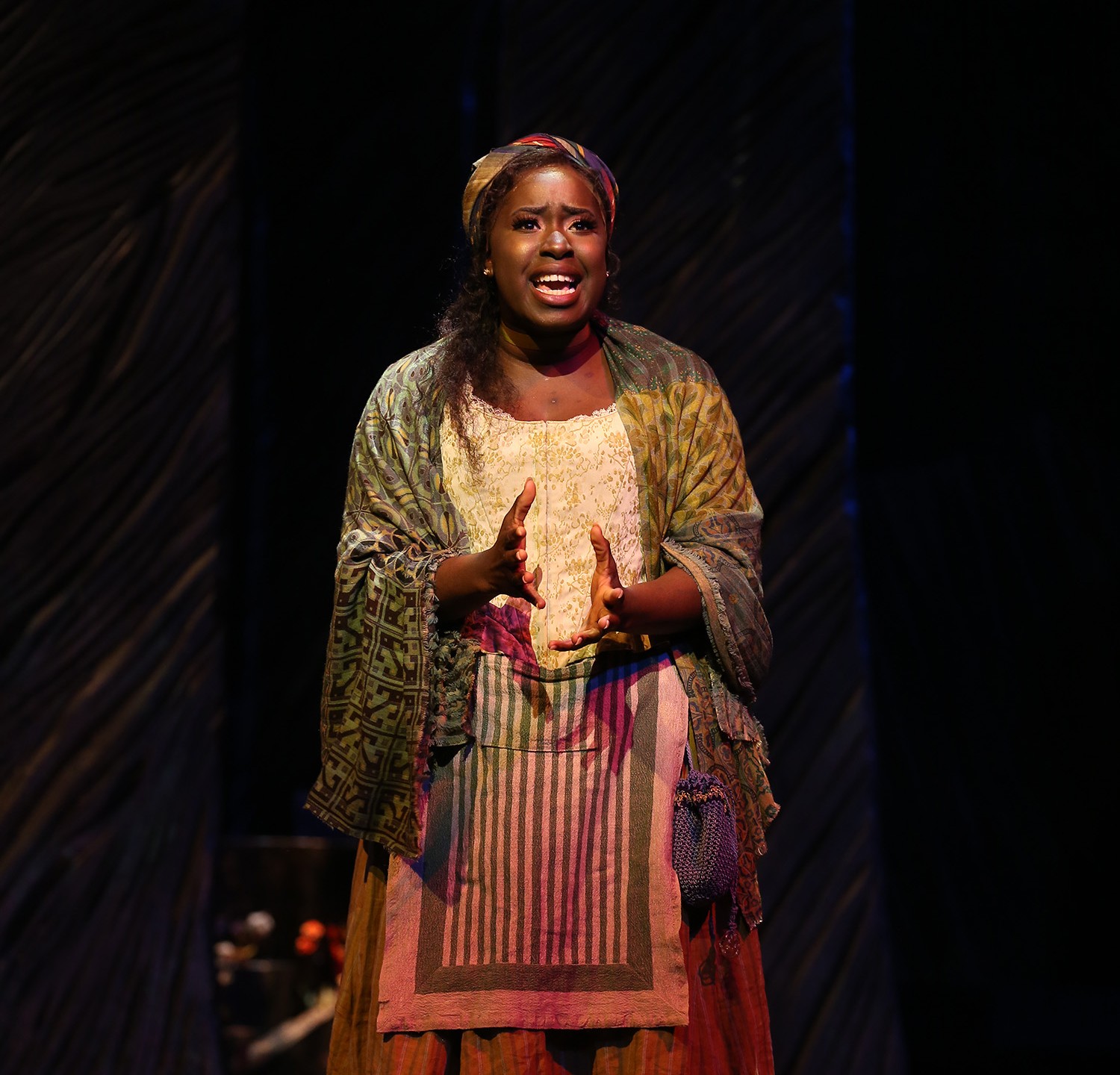 Baker’s Wife stands and sings. She wears a corseted top, a brightly patterned scarf of reds, blues and yellows on her head, and an ankle-length yellow skirt