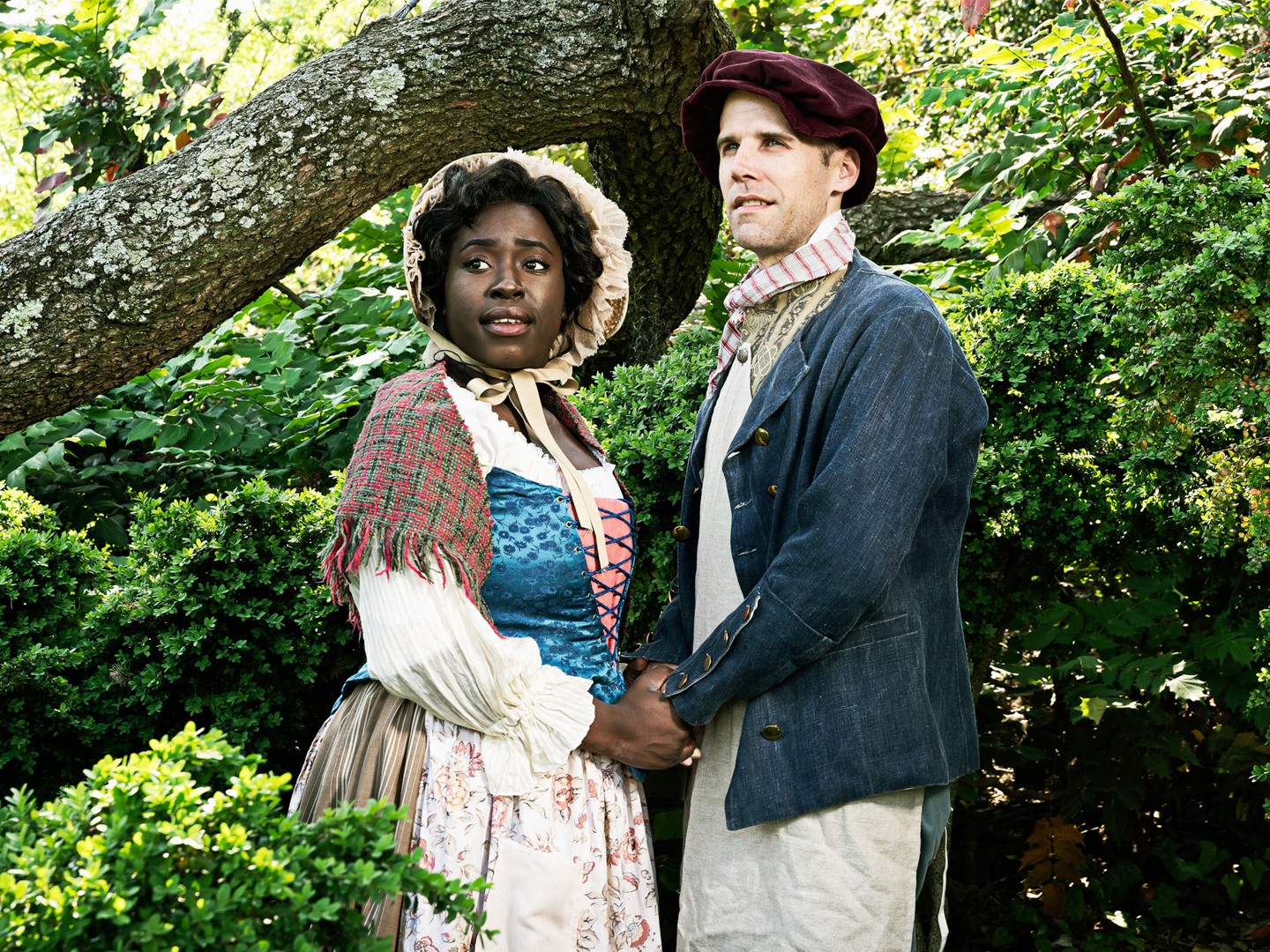 A black woman and white man hold hands in a forest. They are dressed like poor 18th-century fairy tale characters.