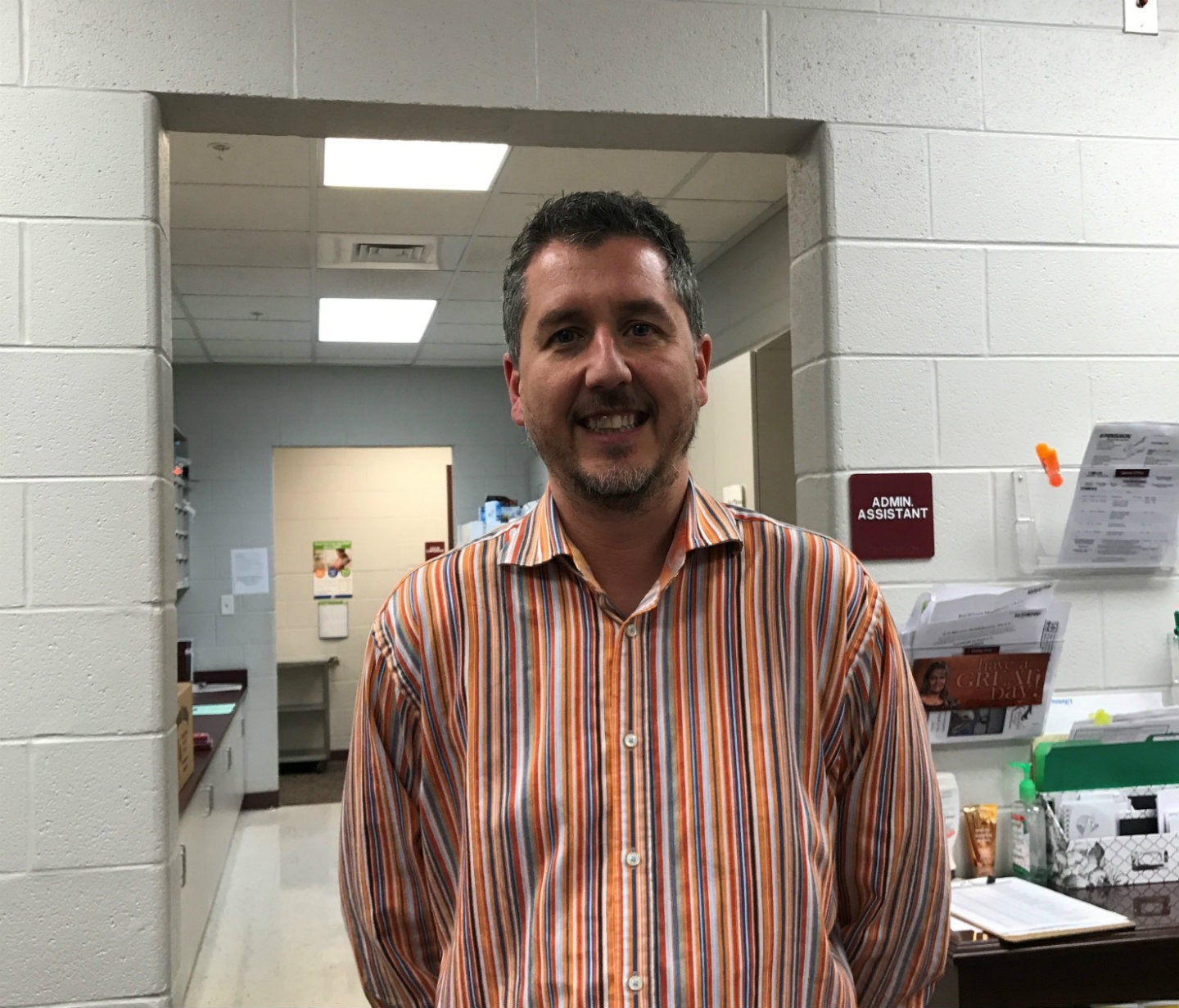 Teacher Joe Moneymaker stands in a doorway at his school. He wears a vertical-striped orange and purple button down shirt.