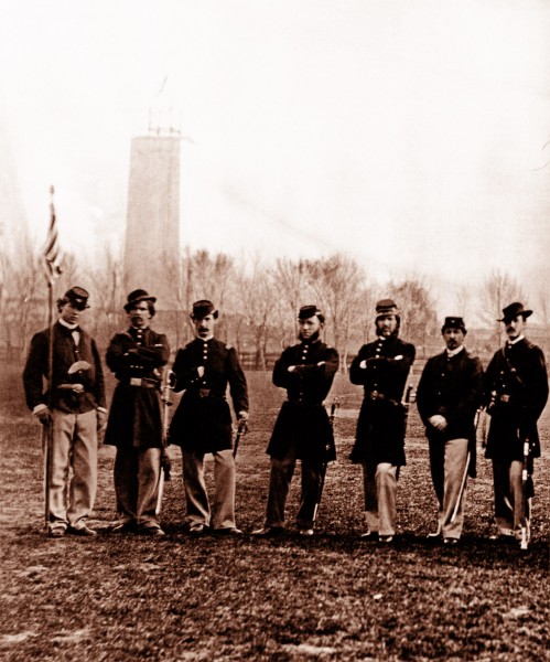 Seven Union soldiers in uniform pose with their muskets and an American flag on the National Mall. Behind them, the unfinished Washington Monument is seen.