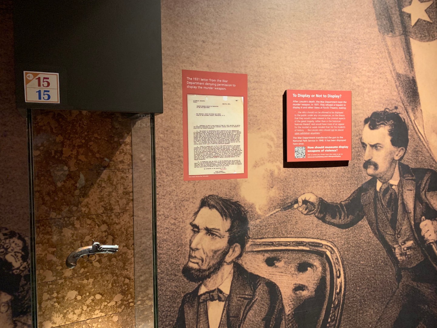 Two museum signs are posted on a wall. One is square sign with text. The other is a rectangular sign with text and an image of a typed letter. Signs are displayed next to a small pistol suspended in a museum display case, and an illustrated mural of assassin John Wilkes Booth shooting President Abraham Lincoln. 