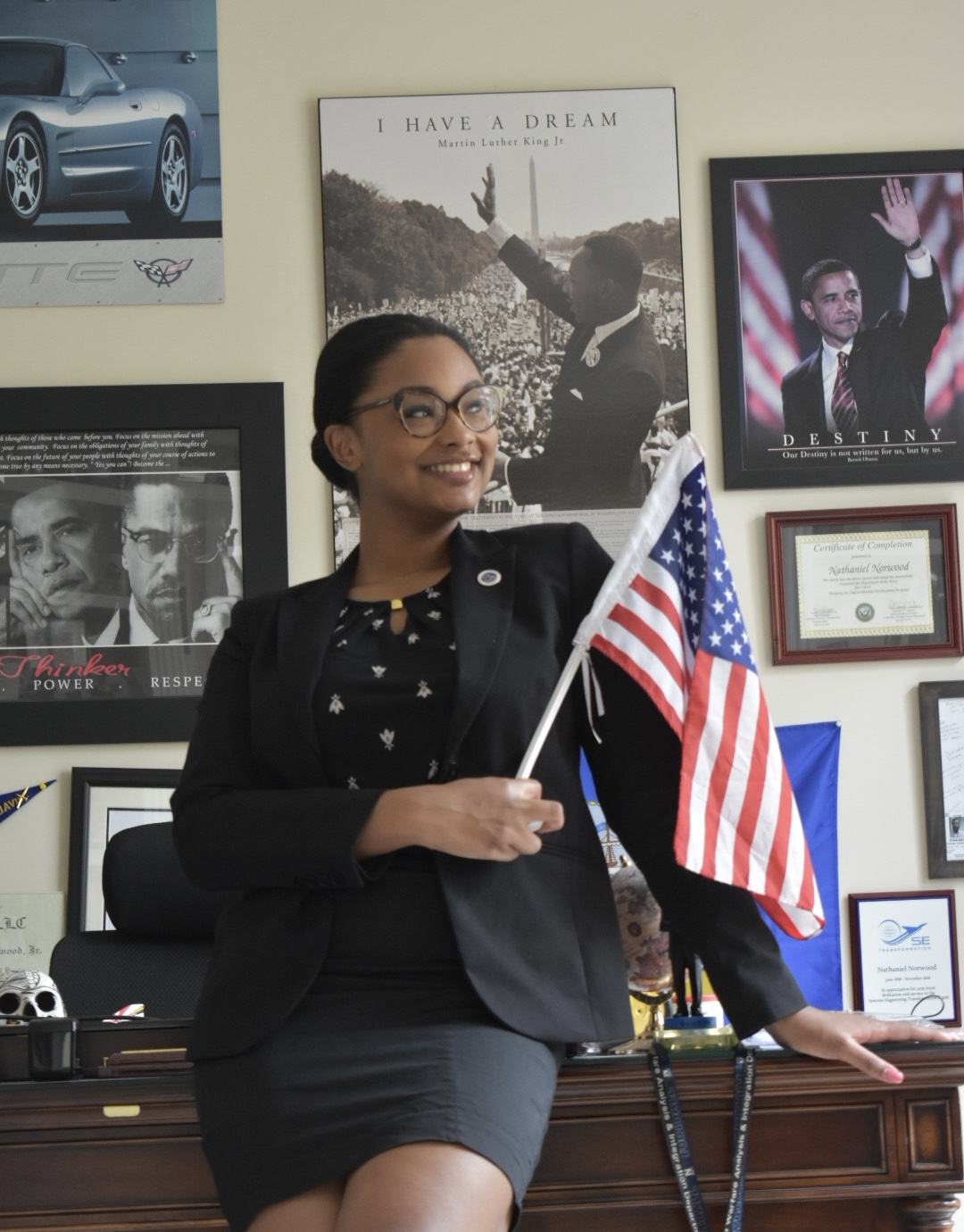 Young woman in a business suit leans near a desk holding a small American flag.