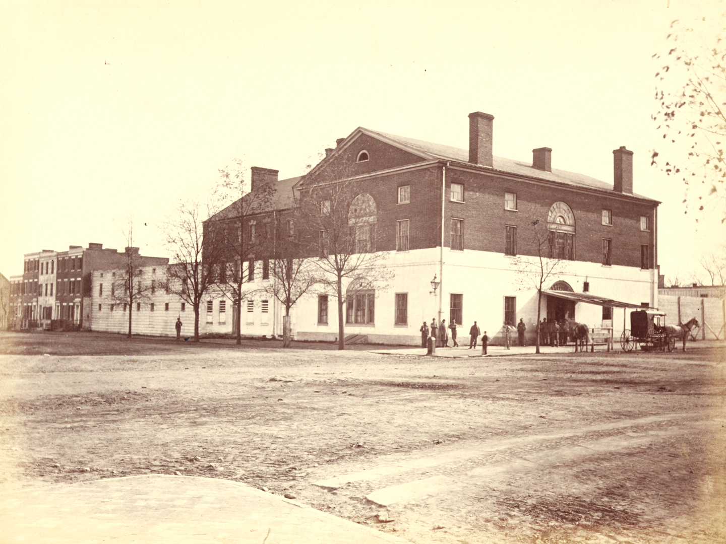 Sepia-toned photograph of three-story brick building with gabled roof and chimneys. Bottom half of building painted white, rest of building dark brick. Awning at front entrance, with people standing around front door.
