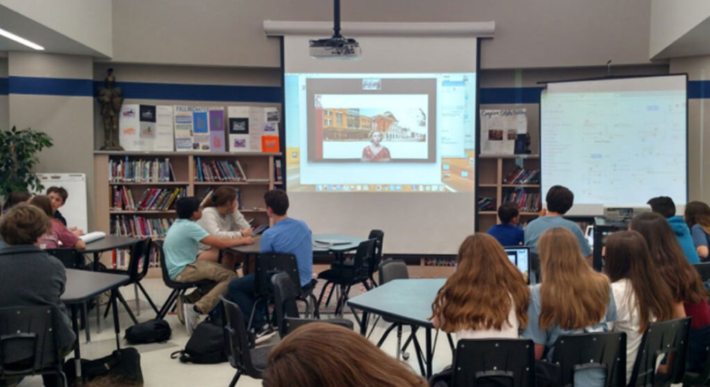 Students gather in a school library to connect with Ford’s Education Staff via videoconferencing. Students are grouped at tables, watching a large projection screen.