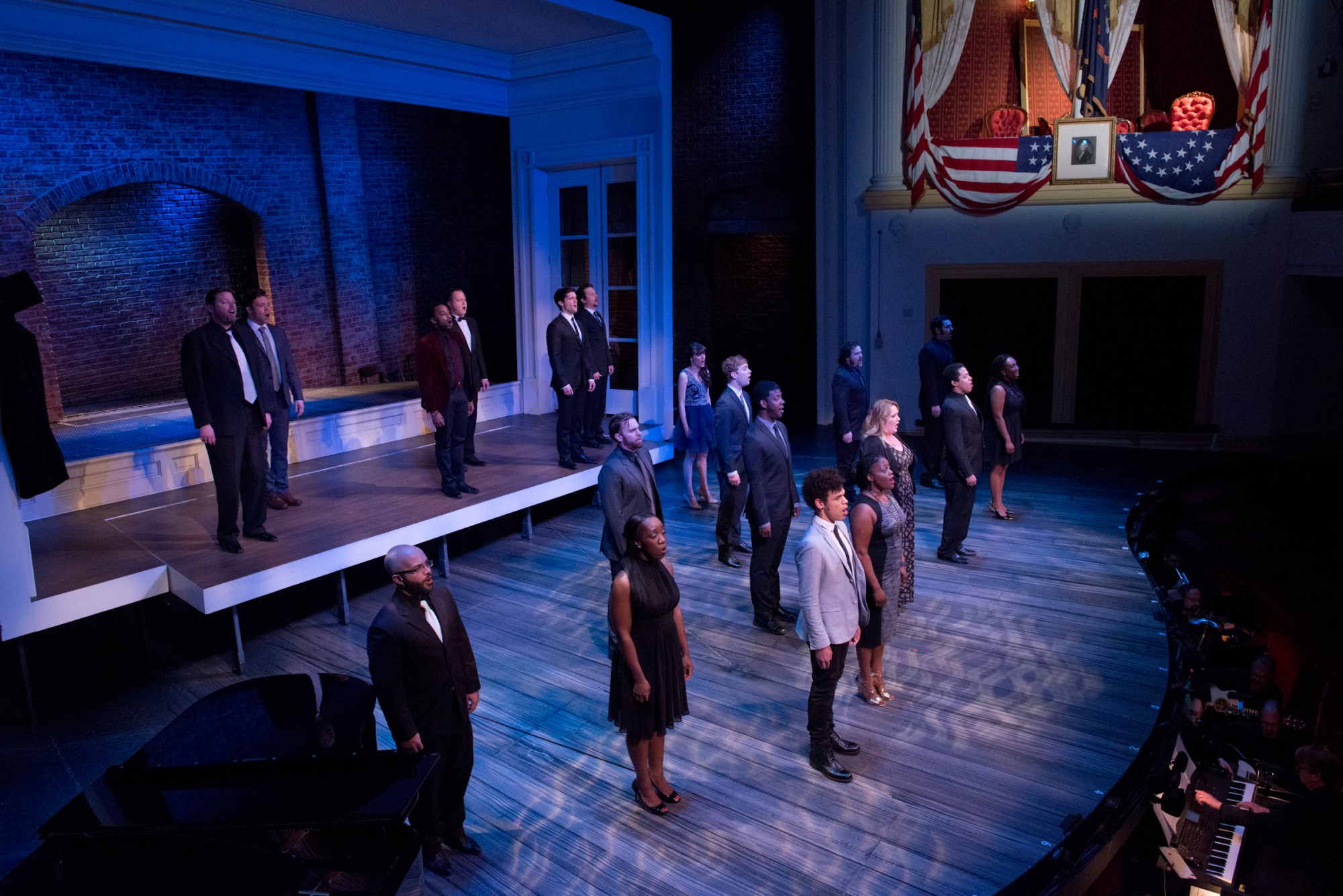 A dozen actors in modern-day street clothes stand in staggered positions on the Ford's Theatre stage. They are illuminated with a moody blue light as they look out toward the hundreds of people seated in the audience. Above the actors on stage and to the right is the Presidential Box, draped with yellow curtains and it's white balcony draped with flags-- just as it was the night of Lincoln's assassination. 