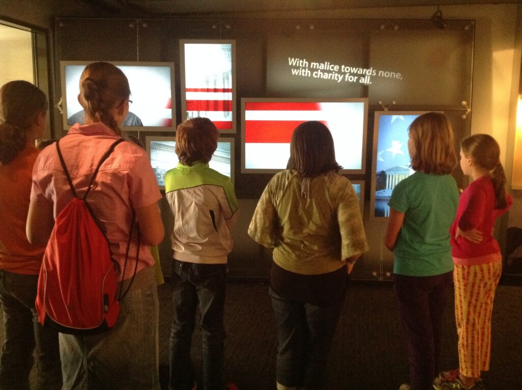 A row of students stand facing away from the camera, looking at a museum display composed of television screens.