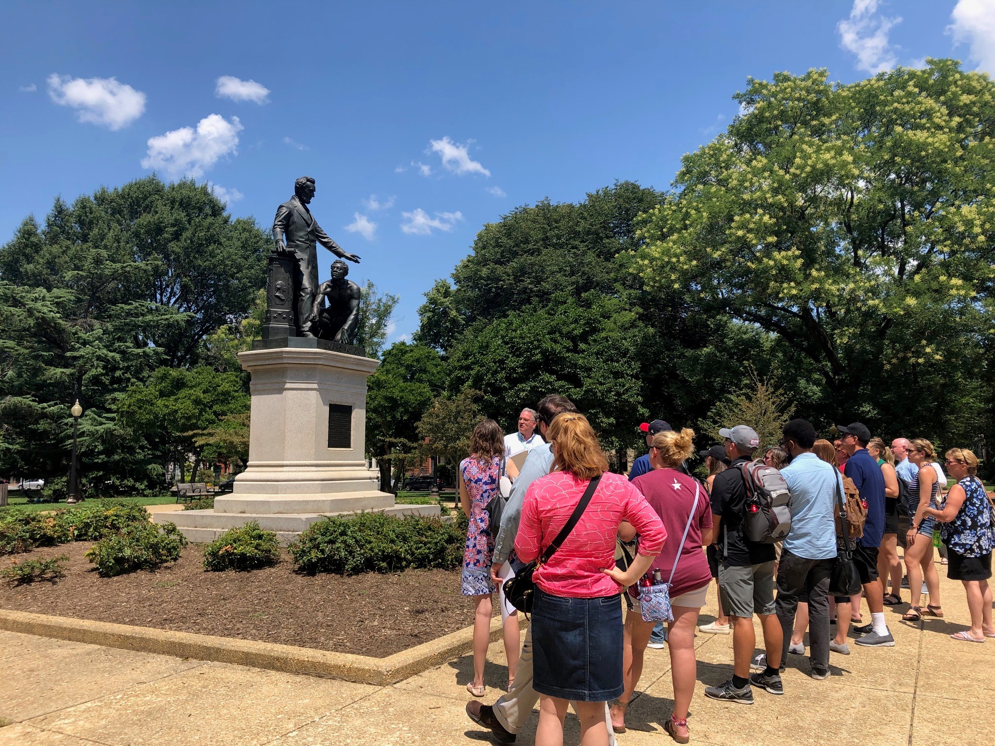 Teachers stand on DC's Lincoln's Park observing a statue of Abraham Lincoln and an emancipated slave. Lincoln stands and the emancipated man kneels beside him.