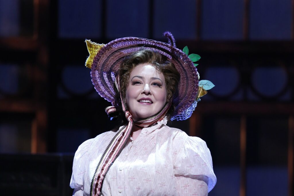 A woman in a light purple dress and a floral purple hat stands on a stage.