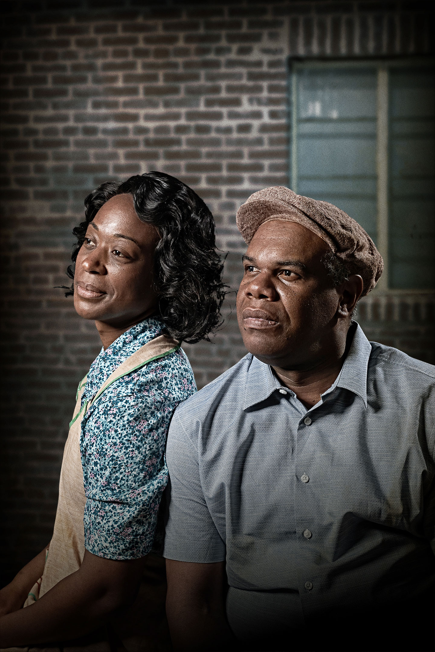 Actors playing August Wilson’s characters Rose and Troy from the play “Fences” sit together in their 1950s costumes. Rose wears a tan apron over a short-sleeved house dress that is patterned with small blue flowers. Troy wears khakis, a short-sleeved blue button-down shirt and a tan linen cap.