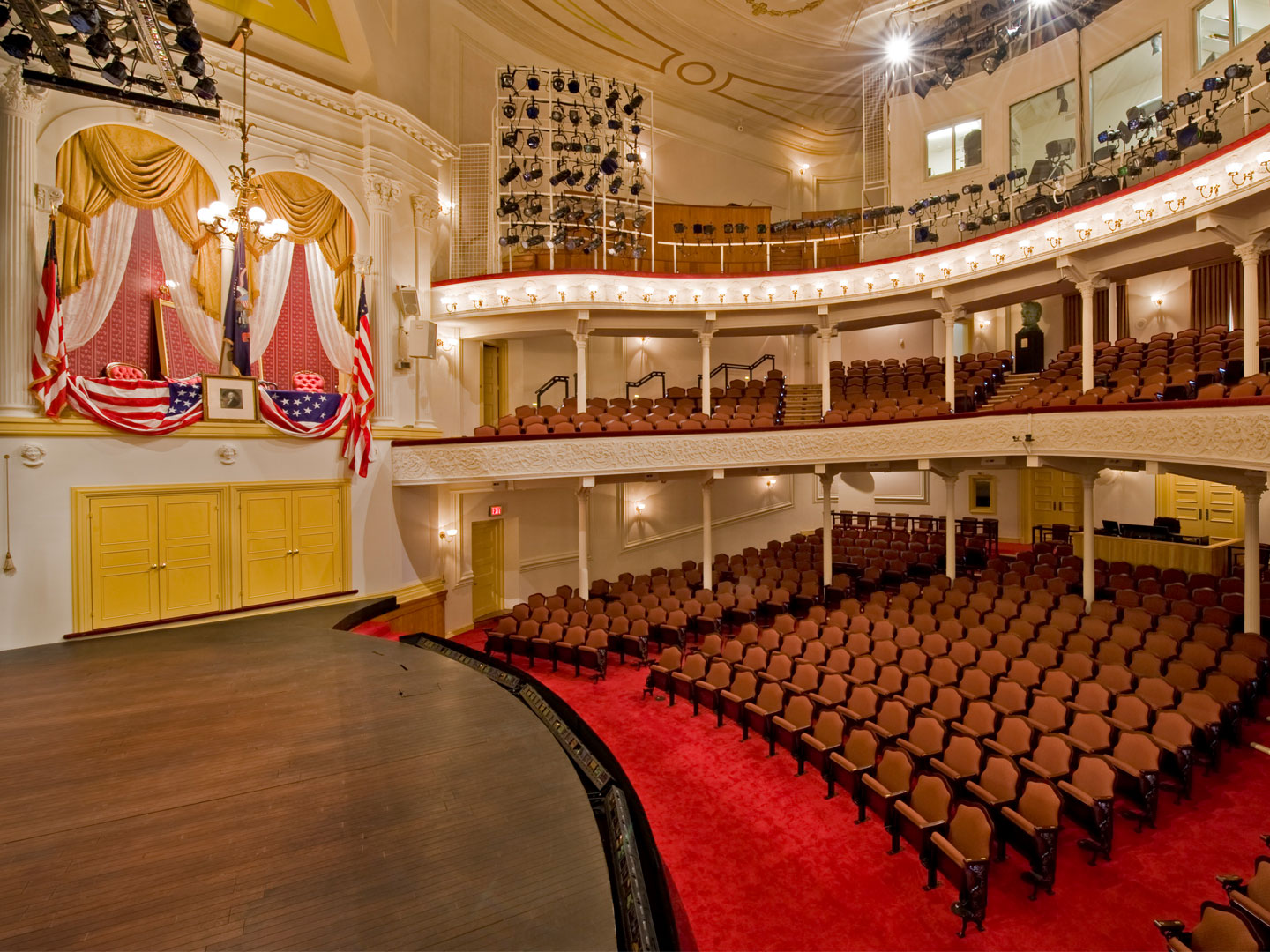 A side view of the stage and seating at Ford’s Theatre. On the left is the President Box with an American flag, a framed picture of George Washington and American flag bunting draped over the box.