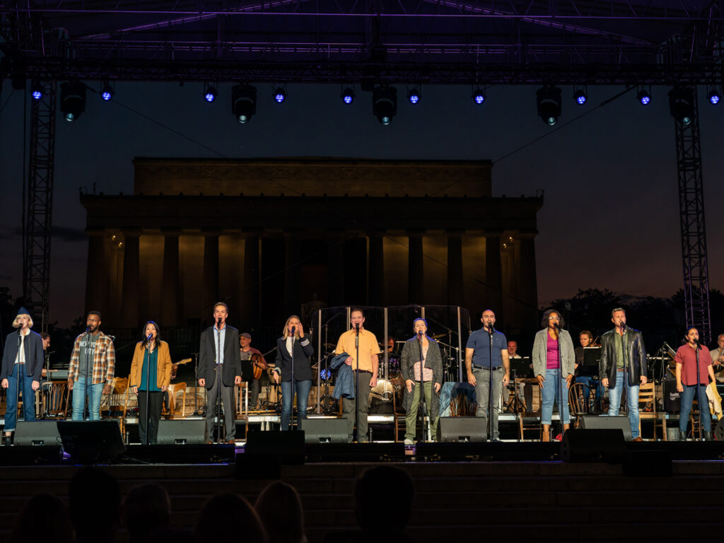 Twelve actors stand on an outdoor stage built near the Lincoln Memorial. Behind the actors sit a band playing drums and Celtic instruments.