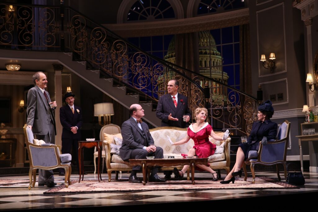 On a set designed to look like a fancy hotel living room in the 1950s, a woman in a red dress and a man in a three-piece suit sit on a sofa. Four others stand around them.