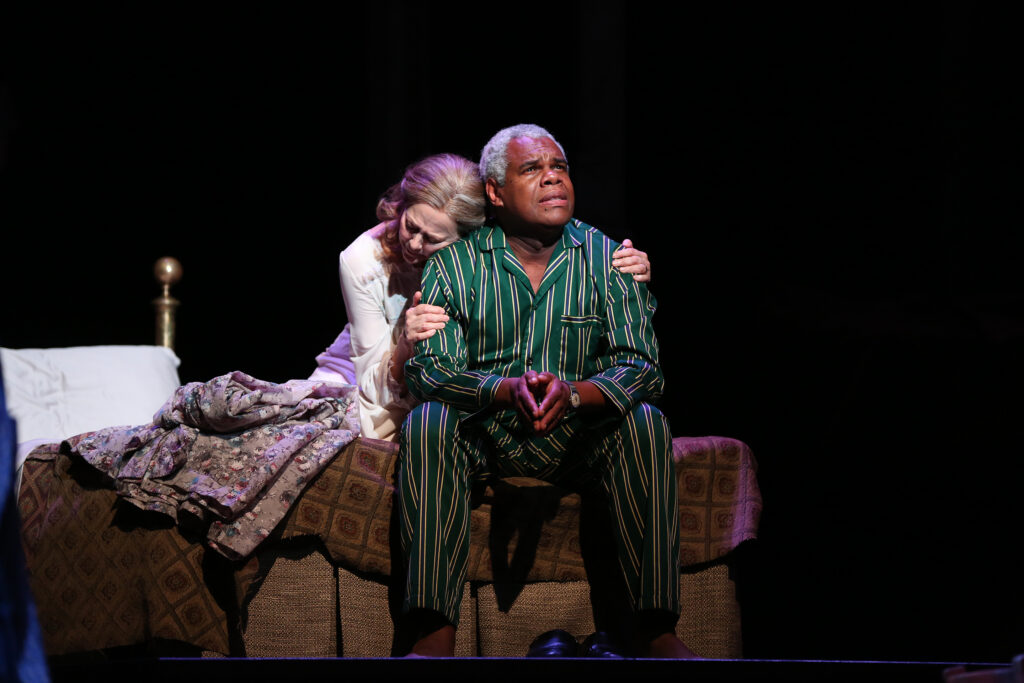 An older woman in a nightgown and robe wraps her arms around the shoulders of her husband, an older man in green and black pin-stripe pajamas. He looks up wistfully.