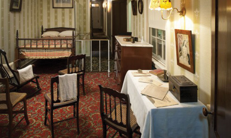 A small bedroom decorated with 1860s-era furniture. The room has green-and-white-striped wallpaper. In the right corner sits a double-sized mattress on a wooden bed frame with two pillows and a colorful quilt with geometric shapes. Small wooden chairs are arranged facing the bed.