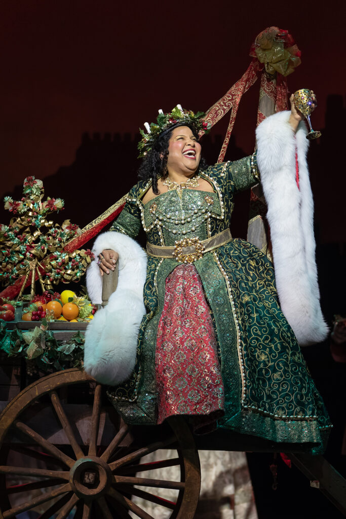 A woman dressed in an embroidered green and red dress sets on the edge of a wagon filled with fruit. She wears a wreath on her head and holds up a goblet of wine while smiling.