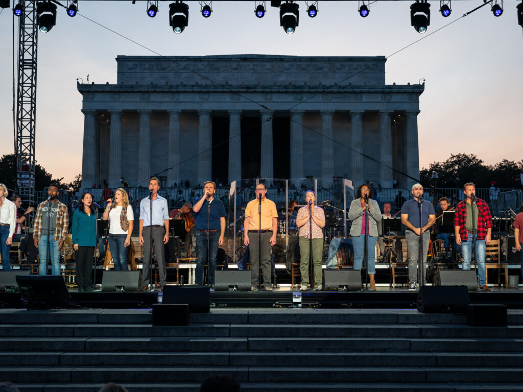 Twelve actors stand on an outdoor stage built near the Lincoln Memorial. Behind the actors sit a band playing drums and Celtic instruments.