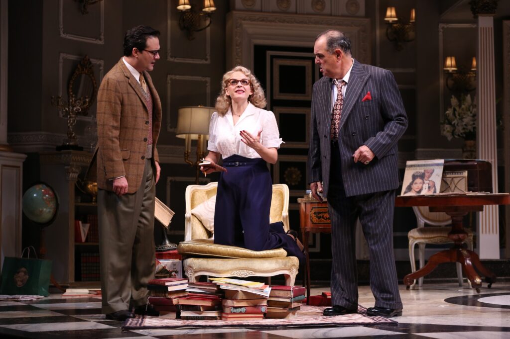 Two men talk to a woman, all dressed in 1950's outfits. The woman kneels on a chair with a large pile of books in front of her.