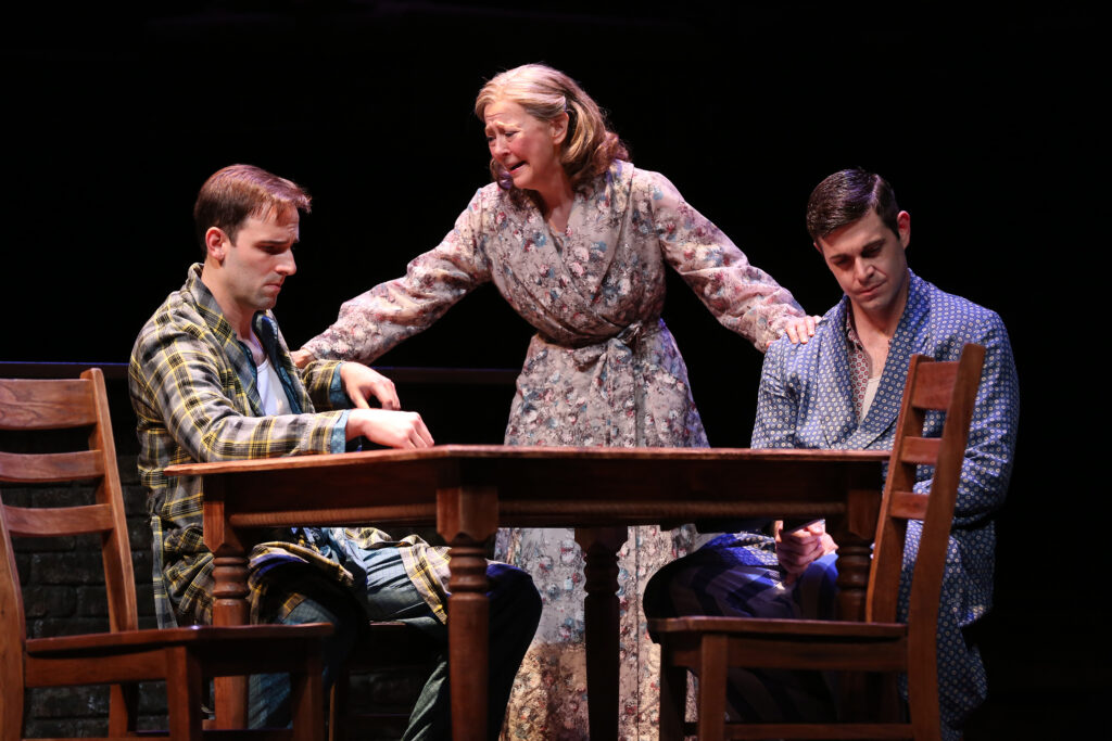 An older woman stands, grasping the arms of her two adult sons, who are seated at a table. Her face is full of pain and pleading. One son looks dismayed. The other looks angry. They are all in their pajamas.