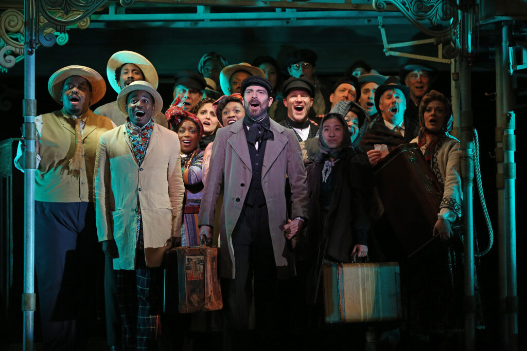 A large group of people in travelling outfits stand together under scaffolding and sing.