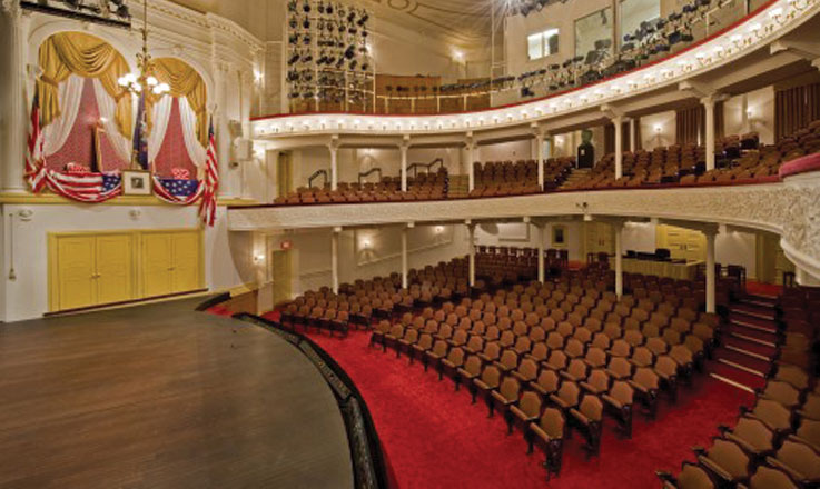 A side view of the stage and seating at Ford’s Theatre. On the left is the Presidential Box with an American flag, a framed picture of George Washington and American flag bunting draped over the box.