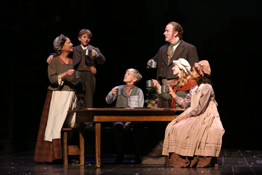 Mrs. Cratchit wraps her arm around Tiny Tim as he makes a toast. Their other family members sit around a table and raise their glasses.