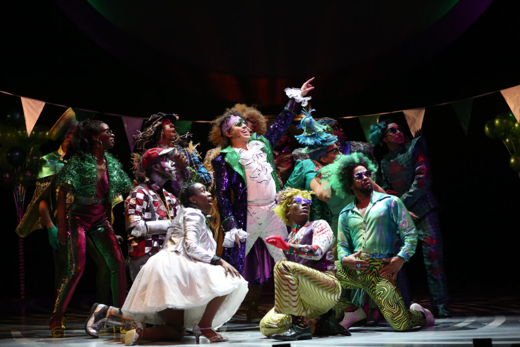The Wiz points to the sky. He wears a white shirt with a ruffle, white pants and a purple-sequin jacket styled after Prince. He is surrounded by people in colorful pants, hats, dresses and wigs.